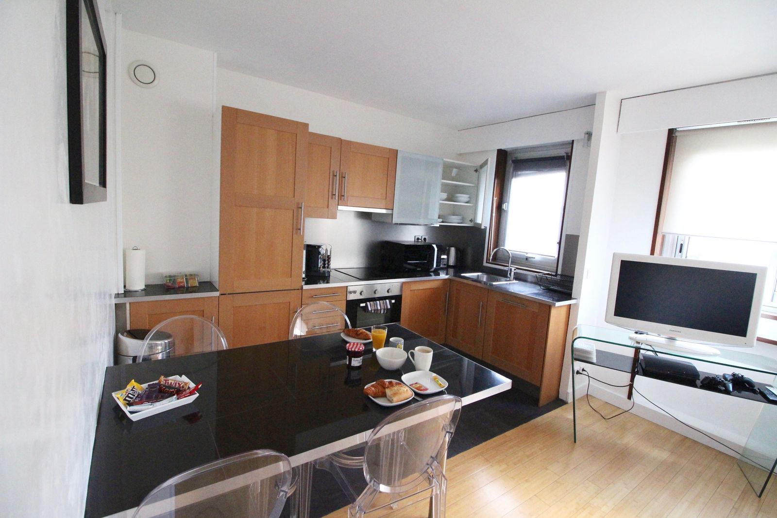 Kitchen-dining area with warm wood cabinets, black dining table, and transparent ghost chairs
